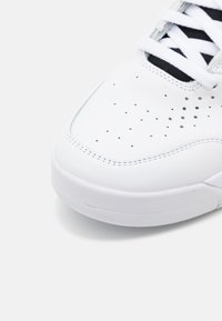 White leather sneaker featuring perforated detailing, textured surface, and flat laces. The sole is rounded with a smooth finish.