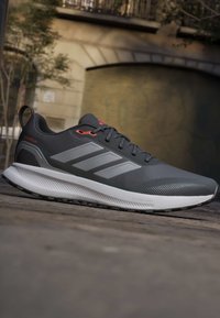 Γκρι και μαύρο παπούτσι για τρέξιμο Adidas με λευκή σόλα και τρεις αντανακλαστικές ρίγες, τοποθετημένο έξω σε πλακόστρωτη επιφάνεια κοντά σε κτίριο.