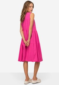 Abito senza maniche fucsia con design a strati, lunghezza ginocchio, realizzato in tessuto leggero. La modella sta in piedi con le mani incrociate dietro la schiena.