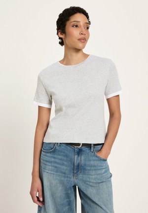 Frau mit kurzen lockigen Haaren, die ein hellgraues kurzärmliges Shirt und blaue Jeans trägt, mit einer Hand in der Tasche vor schlichtem Hintergrund.