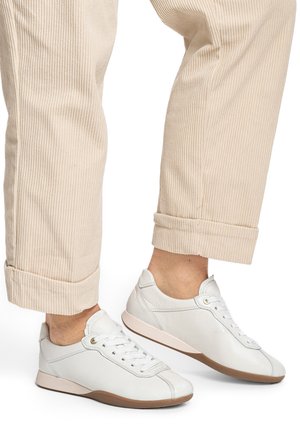 White leather sneakers with a low profile, featuring round laces and a beige rubber sole. Worn with rolled light beige corduroy pants.