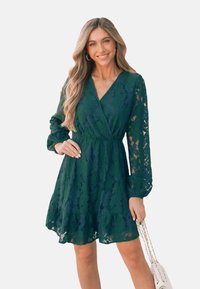 Abito verde in pizzo con maniche lunghe, scollatura a V, vita cintrata e gonna svasata. Presenta un intricato motivo floreale e un orlo stratificato.
