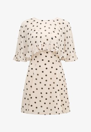 Kremna obleka s črnimi pikami, ki ima nagubano prevleko na prsih in kratke, lahkotne rokave z nagubanimi detajli.