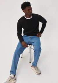 Black long-sleeve shirt with white neckline, light blue jeans, and gray sneakers. Seated on a silver stool against a white background.