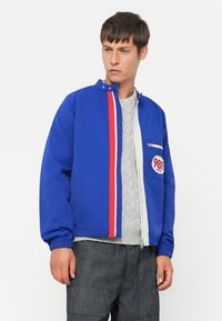Ung mand iført en blå lynlåsjakke med røde og hvide striber og et "981" patch, en grå kabelstrikket sweater og mørkegrå bukser.