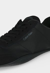Μαύρο αθλητικό παπούτσι Calvin Klein με κορδόνια, χαμηλό προφίλ, λείο επάνω μέρος και υφασμένο πάτο, εμφανιζόμενο από πλαϊνή γωνία.