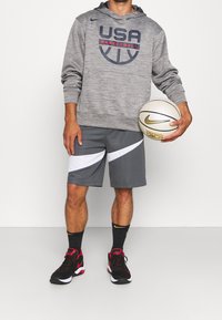 Grau Hoodie mit "USA BASKETBALL" Logo, graue Shorts mit weißen Akzenten, schwarz-rote Basketballschuhe, hält einen Nike Basketball.