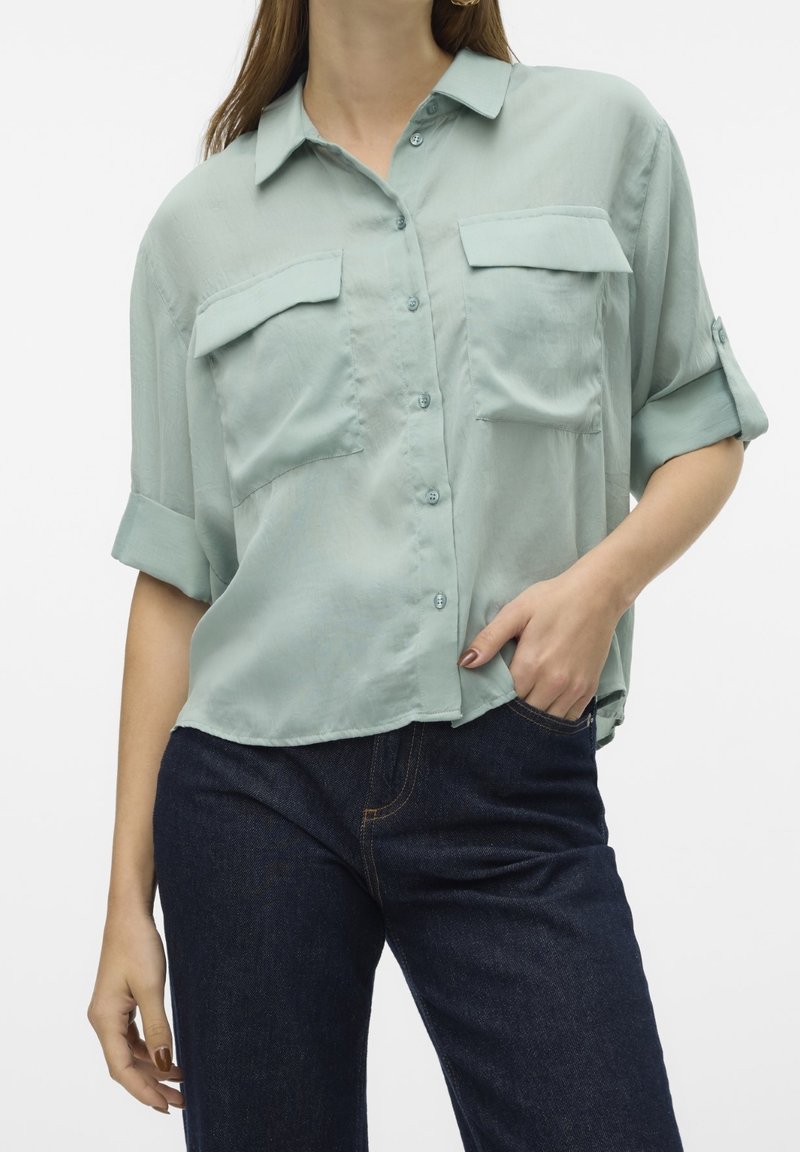 Femme portant une chemise ample vert clair boutonnée avec les manches retroussées et un jean bleu foncé, une main dans la poche.