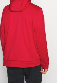 Personne portant un sweat à capuche rouge avec la capuche baissée et un pantalon noir, vue de dos sur un fond clair.