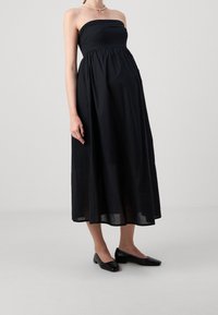 Robe noire sans bretelles avec un corsage ajusté et une jupe fluide mi-longue. Portée avec des ballerines noires texturées. Tissu lisse, design simple.