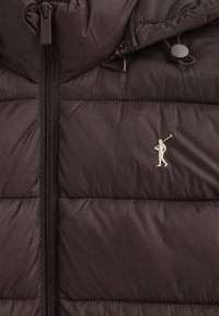 Brun vadderad jacka i slätt, mjukt material med vertikal dragkedja och en logotyp broderad på bröstet. Designad med huva.