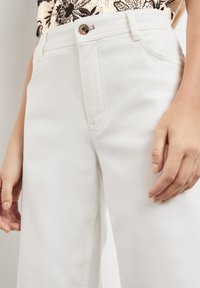 Pantalones de mezclilla blancos con una cintura ajustada, cierre de botón de metal, bolsillos laterales y una tela texturizada, mostrados desde la cintura hasta la rodilla.