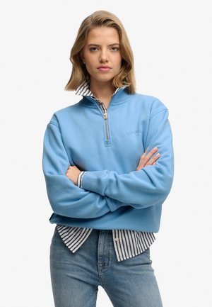 Woman with folded arms wearing a light blue quarter-zip sweatshirt over a black-and-white striped button-up shirt and blue jeans.