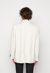 Blazer bianco con vestibilità su misura, rever a taglio, spacchetto singolo sul retro e polsini con bottoni. Texture liscia con un design minimalista.