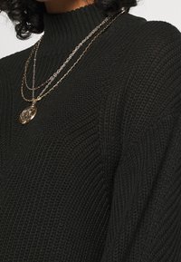 Svart stickad tröja med hög krage, med texturerad tyg. Lager av guldkedjor med en dekorativ hängsmycke bärs över tröjan.