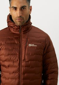 Brown puffer jacket with quilted texture, high collar, and front zipper. Features Jack Wolfskin logo on the left chest. Suitable for cold weather.