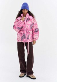 Kurtka puchowa w różowym kolorze z kwiatowymi wzorami, z wysokim kołnierzem, frontowym zamkiem błyskawicznym i dwoma kieszeniami. Połączona z brązowymi spodniami o szerokich nogawkach i beżowymi botkami.