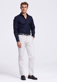 Marine blouse met knopen, witte broek, zwarte leren schoenen. Het shirt heeft een zachte textuur, een op maat gemaakte pasvorm en lange mouwen. Eenvoudige riem als accent.