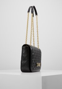 Svart vadderad läderhandväska med guldkedjor och en gyllene metallplakett på en vit pedestal.