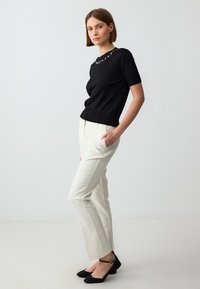 Black short-sleeve top with embellished neckline, paired with cream tailored pants. Model wearing black flats, standing against a neutral background.