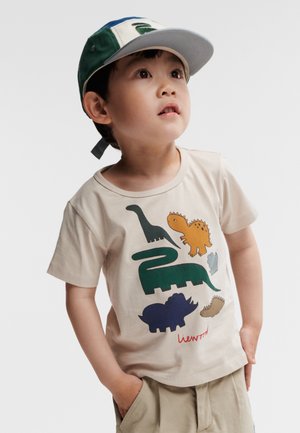 T-shirt en coton beige avec des graphismes de dinosaures colorés, et un logo rouge en bas. Col rond, manches courtes.