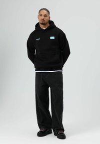 Sweat à capuche noir avec poche avant, arborant un logo bleu. Associé à un jean foncé ample et des chaussures noires épaisses. Fond neutre.