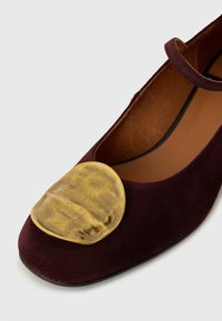 Scarpa in suede bordeaux con punta arrotondata, caratterizzata da una grande accentuazione metallica dorata sul davanti. Fodera interna in pelle marrone.