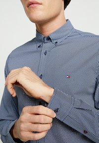 Uomo che abbottona il polsino di una camicia a bottoni blu e bianca con un piccolo logo sul petto.