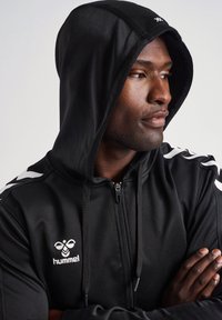 Sudadera con cremallera negra y capucha con cordón, con franjas laterales blancas y un logo en el pecho. Fabricada con una tela atlética suave.
