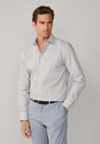 Gestreiftes Langarmhemd in Weiß und Hellgrau, mit Button-Down-Kragen, taillierter Passform und Knopfverschluss. Getragen mit hellgrauen Hosen.