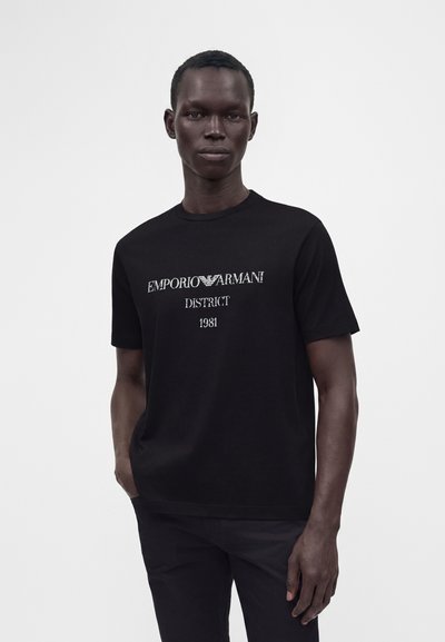 Άνδρας που φοράει μαύρο T-shirt Emporio Armani με λευκά γράμματα που αναγράφουν "EMPORIO ARMANI DISTRICT 1981", στέκεται μπροστά σε απλό λευκό φόντο.