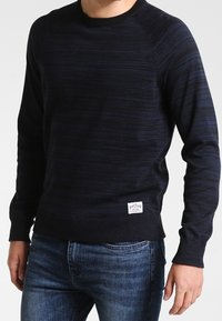 Granatowy sweter z okrągłym dekoltem w paski, z ściągaczami przy mankietach i u dołu, z małą prostokątną metką marki na dolnej lewej stronie.