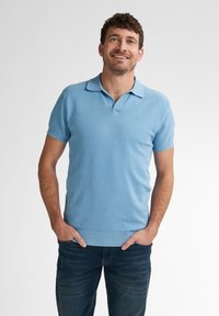 Světle modré polo tričko s texturovaným vzorem, krátké rukávy a žebrový límec, nošeno s tmavě modrými džíny, ruce v kapsách.