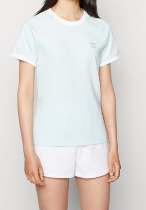 Mujer vistiendo una camiseta de manga corta azul claro de Adidas con rayas blancas y pantalones cortos blancos, de pie con los brazos relajados a los costados.