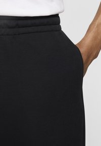 Melnas sporta bikses ar mīkstu tekstūru, kurām ir pieguļošs dizains un elastīga josta. Tuvplāns uz jostas un sānu šuvju detaļām.