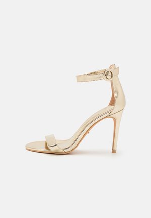 Guld højhælet sandal med ankelsnor og rund spænde, åben tå og slank stilethæl på en ensartet hvid baggrund.