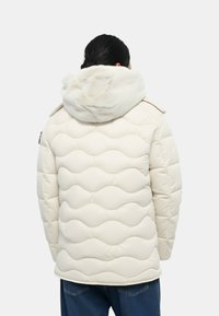 Chaqueta acolchada de color crema con un patrón de olas texturizado y capucha de piel sintética. Cuenta con cierre de cremallera y se lleva sobre unos vaqueros.