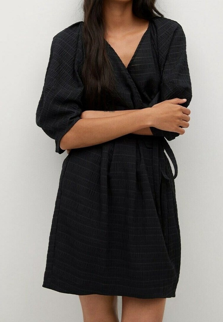 Femme aux longs cheveux foncés portant une robe portefeuille noire texturée avec des manches bouffantes trois-quarts, les bras croisés, debout devant un fond uni.