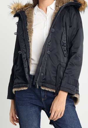 Frau trägt eine schwarze Winterjacke mit Pelzbesatz an Kapuze und Innenfutter, weißes Hemd und blaue Jeans, Hand ruht nahe dem Oberschenkel.