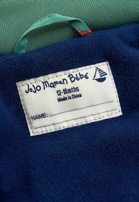 Jūrinio laivyno pūkų audinys su baltu lipduku, kuriame mėlyna spalva užrašytas tekstas, burlaivio grafika ir laukeliai vardui bei amžiui. Žalias tekstūruotas audinys dalinai matomas.