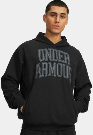 Junger Mann trägt einen schwarzen Kapuzenpullover mit der Aufschrift "UNDER ARMOUR" in großen Buchstaben auf der Brust, die Hände in den Taschen des Kapuzenpullovers.