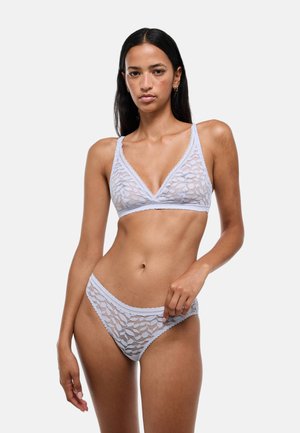 Femme portant un soutien-gorge et une culotte en dentelle gris clair assortis, debout devant un fond clair uni avec une expression neutre.