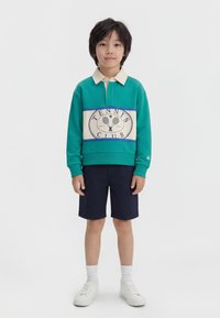 Αγόρι που στέκεται μπροστά, φοράει πετρόλ φούτερ με τη λέξη "Tennis Club", μπλε σκούρα σορτς, λευκές κάλτσες και λευκά αθλητικά παπούτσια, σε απλό φόντο.