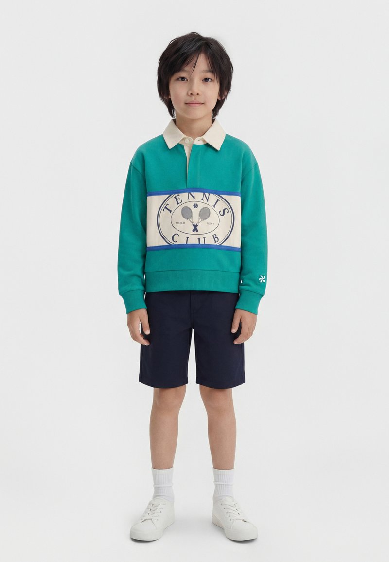 Αγόρι που στέκεται μπροστά, φοράει πετρόλ φούτερ με τη λέξη "Tennis Club", μπλε σκούρα σορτς, λευκές κάλτσες και λευκά αθλητικά παπούτσια, σε απλό φόντο.