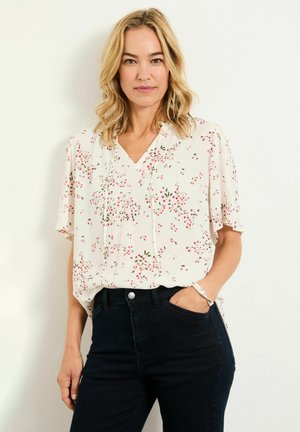 Frau mit schulterlangem blondem Haar, die eine weiße Bluse mit Blumenmuster und dunkle Jeans trägt, mit einer Hand in der Hosentasche vor einer weißen Wand stehend.