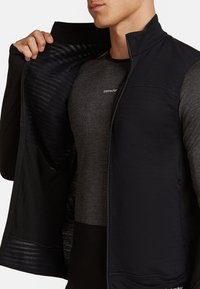 Uomo che indossa un gilet scuro con zip sopra una maglietta grigia a maniche lunghe, tieni il gilet aperto per mostrare l'interno testurizzato.