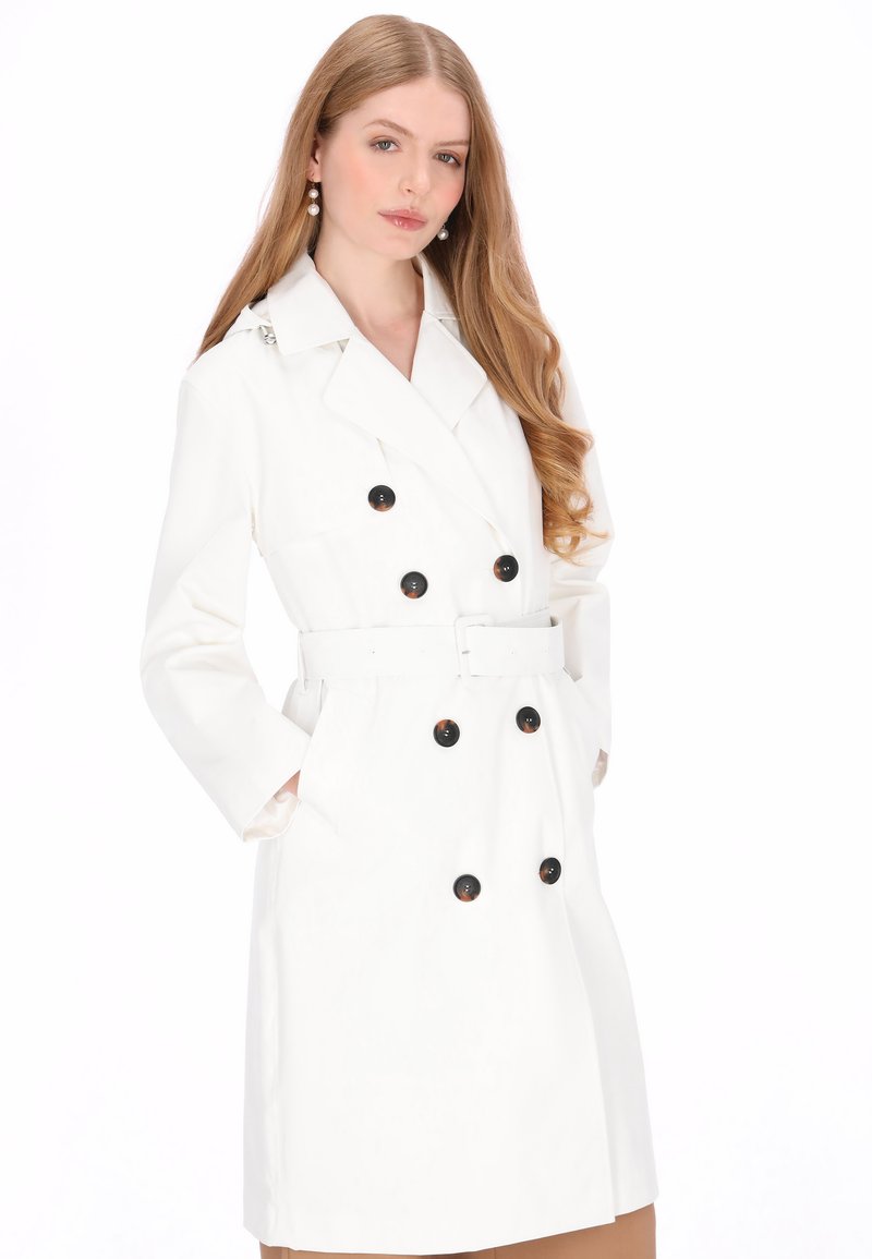 Trenci alb cu design cu nasturi dubli, nasturi negri și talie cu curea. Are mâneci lungi și buzunare laterale.