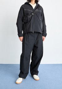 Svart lättviktsjacka med dragkedja och huva, tillsammans med lösa svarta byxor. Inkluderar en liten crossbody-väska och vita sneakers.