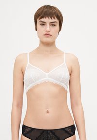 Bralette en dentelle blanche avec des bretelles ajustables, motif floral, bords festonnés et forme triangulaire. Associé à un sous-vêtement en dentelle noire.