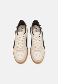 Beige Sneaker mit einem oberen Teil aus glattem Leder, durchlöchtem Zehenbereich, dunkelblauen Seitenakzenten und einer Gummisohle. Die Schnürsenkel sind mit einem strukturierten Muster gewebt.